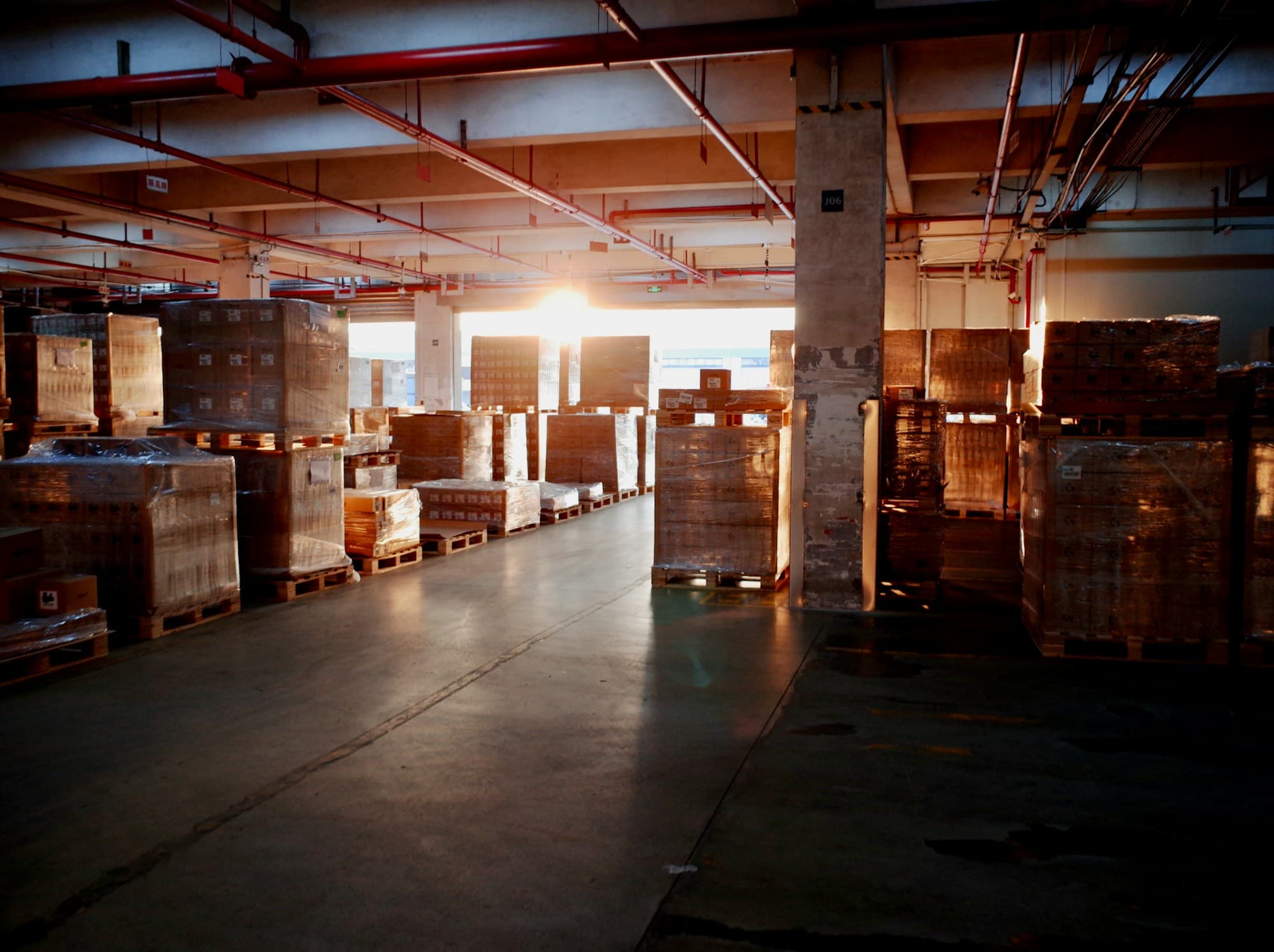 Shrink-wrapped pallets of goods stacked on a warehouse floor, backlit by daylight through open loading doors.